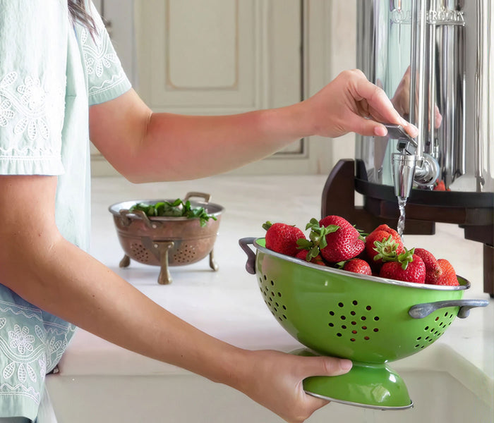 A person in a kitchen uses Clearbrook gravity system to rinse a bright green colander filled with fresh strawberries. 